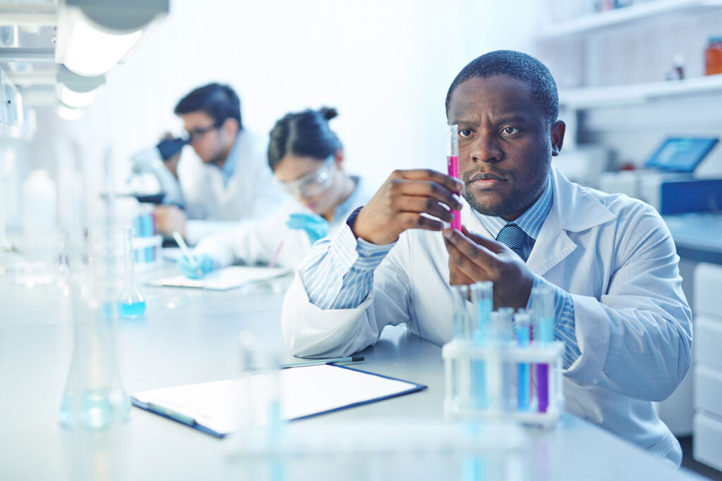African American scientist holding a test tube with pink liquid and two other scientist working in the background