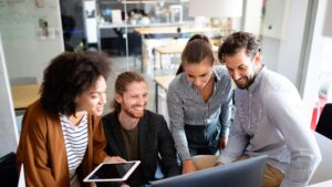 a group of 4 coworkers looking at a laptop screen and one is holding a tablet