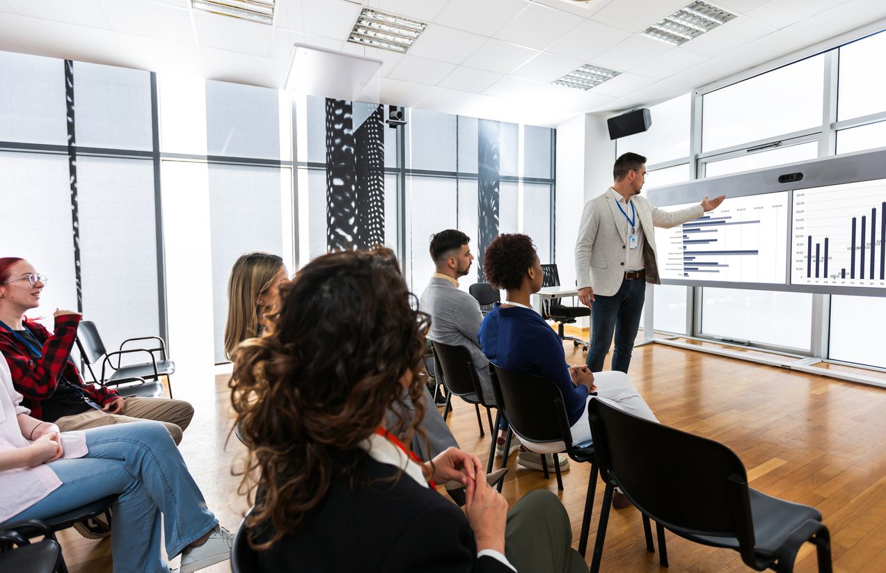 Male CEO standing in front of a multiracial group of colleagues. Multimedia classroom with smart board and graphics on them.