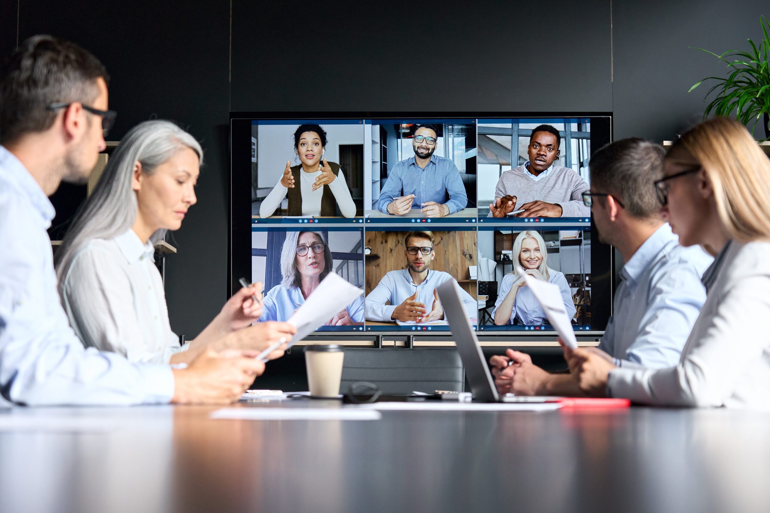 Global corporation online videoconference in meeting room with diverse people sitting in modern office and multicultural multiethnic colleagues on big screen monitor.