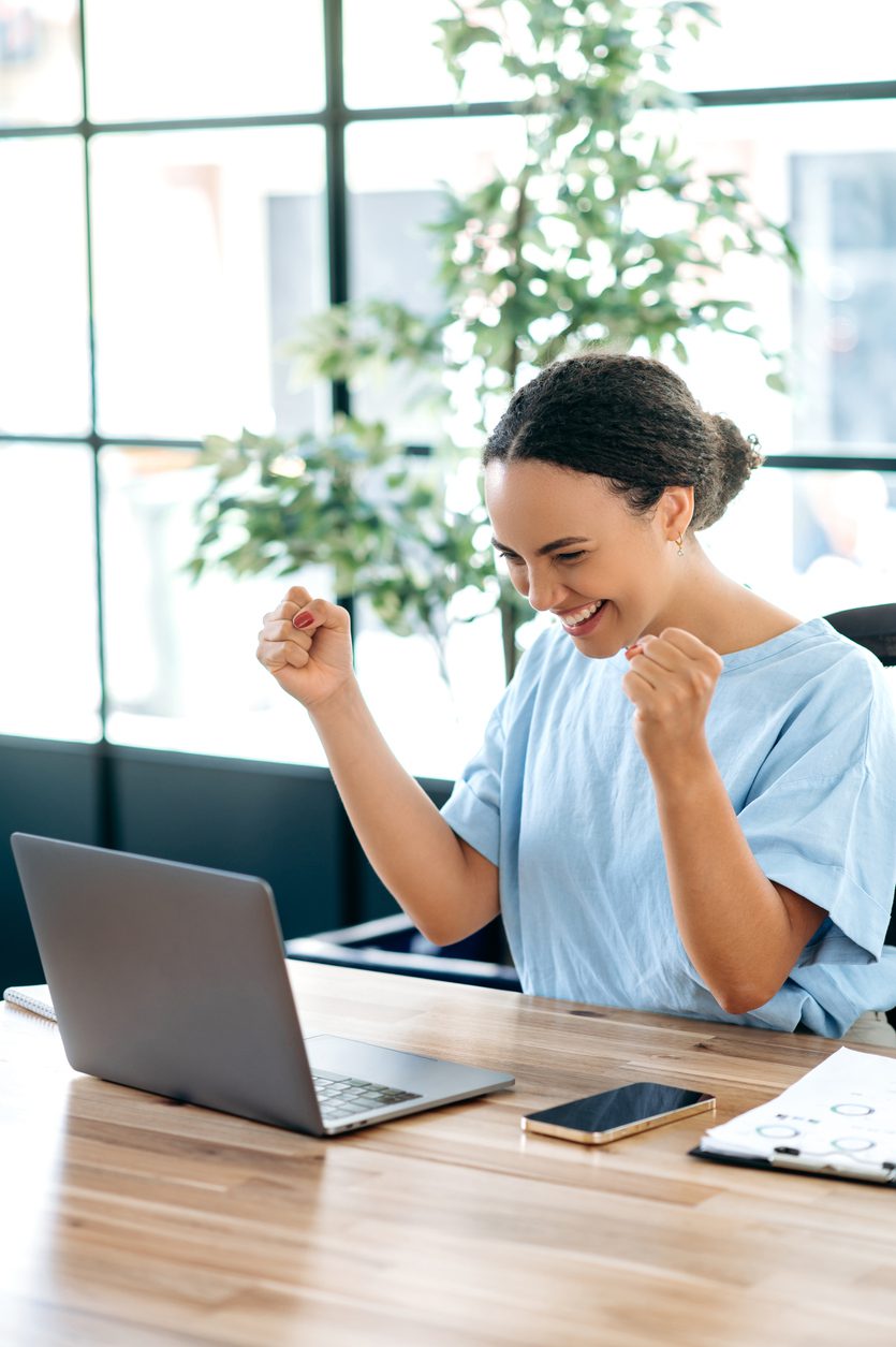 Vertical photo of joyful hispanic or brazilian business woman, sits at a work desk with laptop in office, rejoicing in success, big profit, celebrate deal, gesturing with fists, looks at laptop, smile