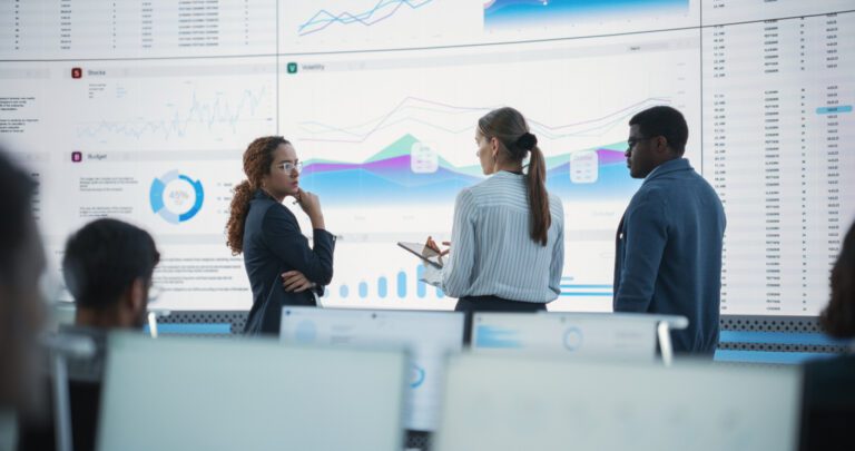 Diverse Team Of Data Scientists Having A Meeting And Discussing Reports In a Modern Monitoring Office With Analytics Feed On a Big Digital Screen. Deep Learning Company With Employees Behind Computers