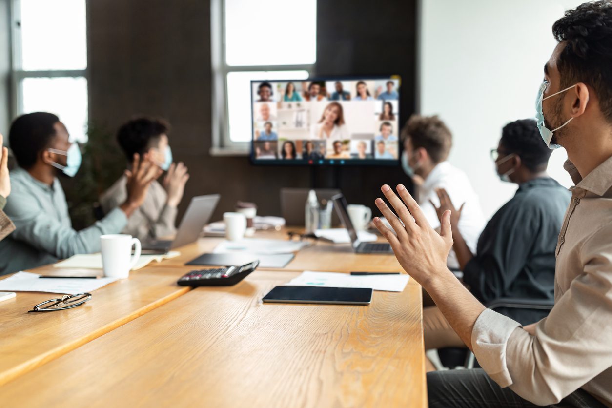 Videoconferencing Concept. Diverse group of workers in face masks making video call with colleagues, sitting at table in boardroom, waving at screen at office, talking on web with remote employees