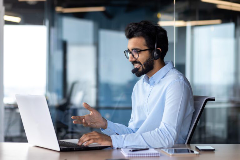 Indian young man wearing a headset conducts a webinar, online conference sitting at a laptop in the office.