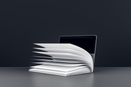 An open book with pages turning, set in front of a laptop on a grey surface. Dark background. Concept of digital learning and education. 3D Rendering